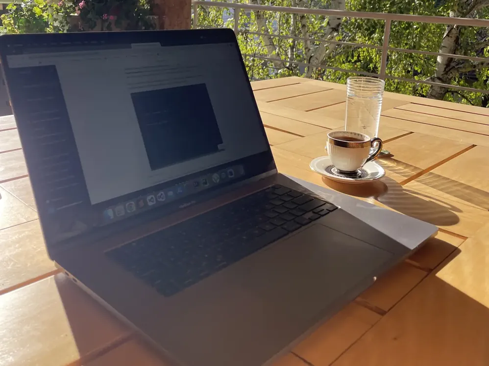 A laptop open on a sunny wooden balcony table, beside a cup of coffee and a glass of water, with leaves behind.