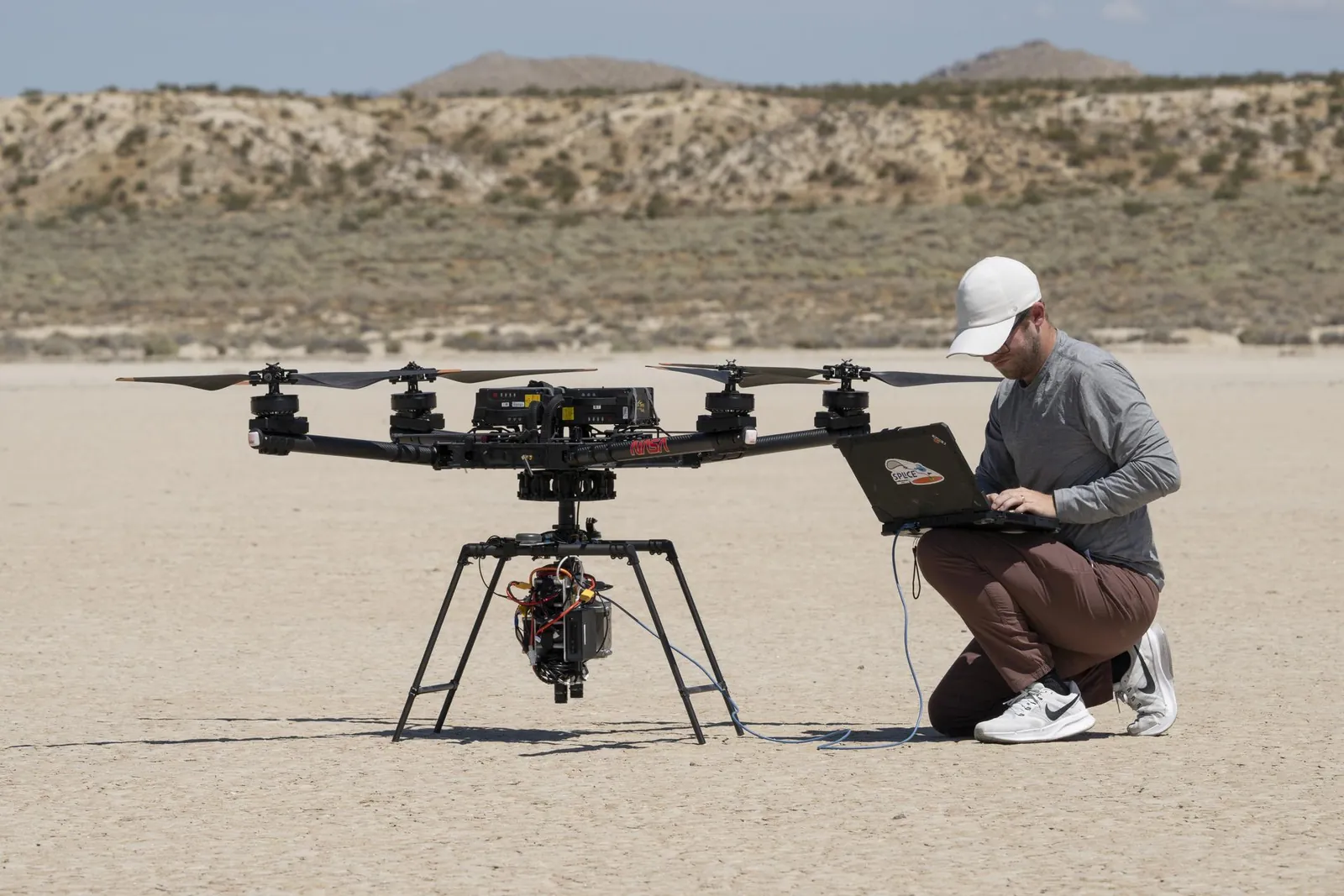 On a dry lakebed, a project manager kneels with a laptop beside a large drone, checking its guidance system.